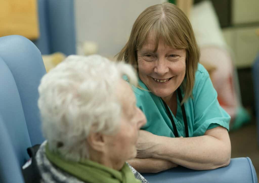 Smiling carer in green scrubs leaning toward an elderly person in a chair, reflecting nursing home care